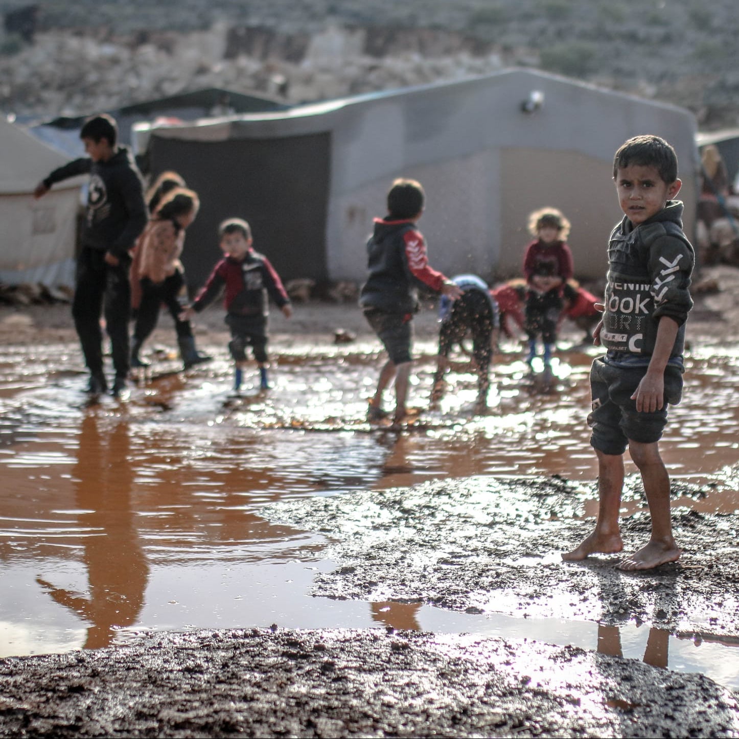 Children playing in muddy water with tents and vehicles in the background, text 'Winter Shelter Appeal Gaza' at the top.